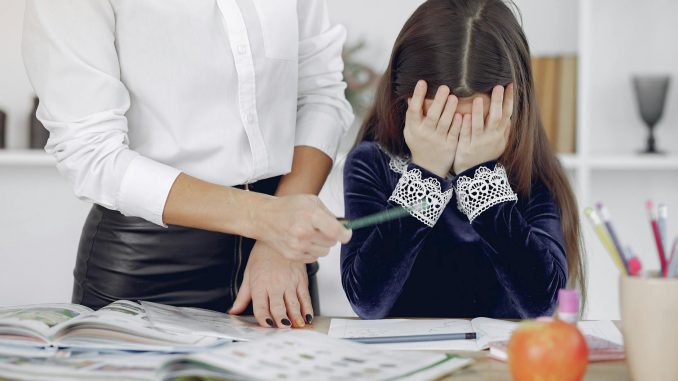 upset little girl sitting near crop woman in classroom